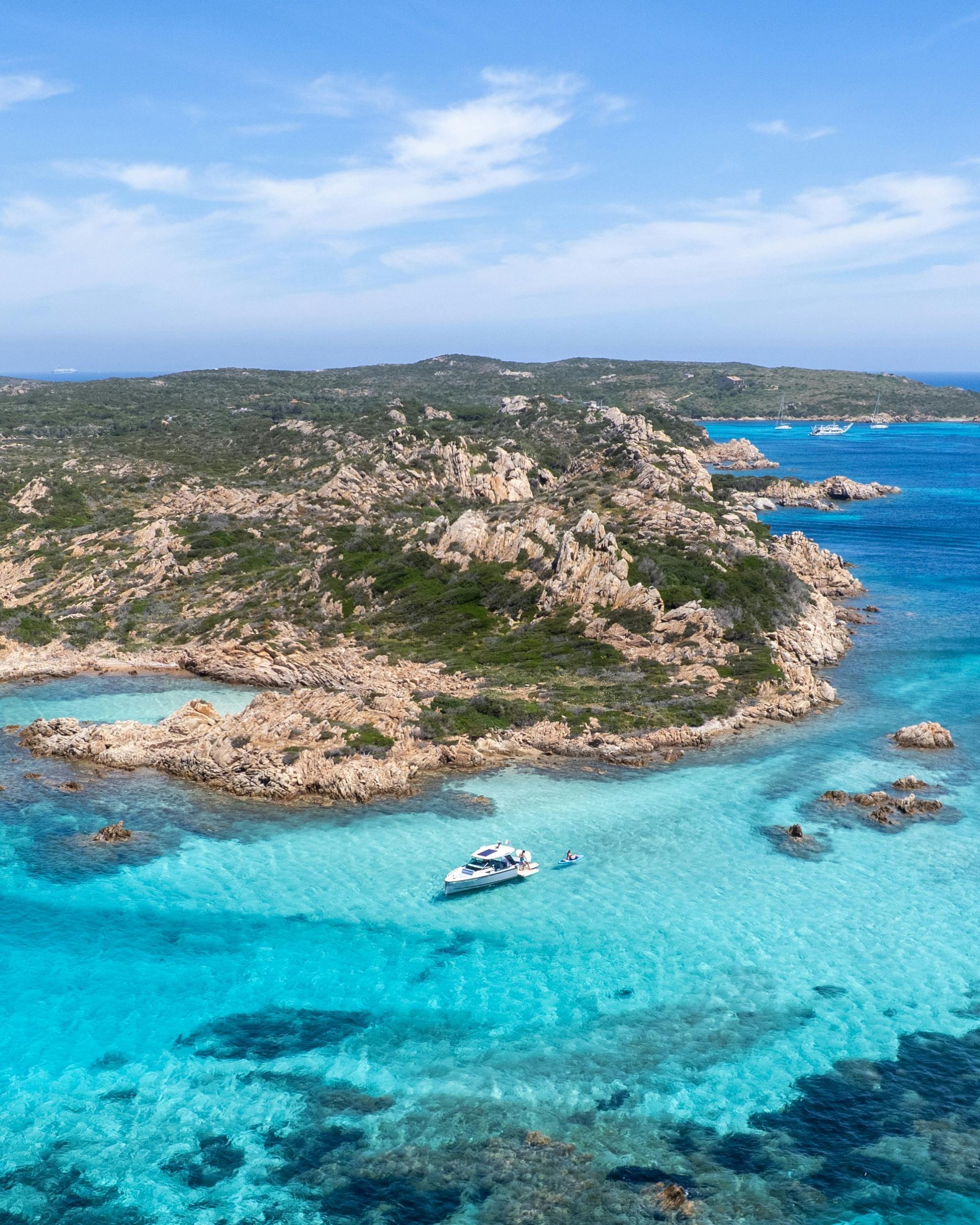 ARCIPELAGO DI LA MADDALENA Aerial shot of a boat in stunning turquoise waters near rocky coastline under blue skies.